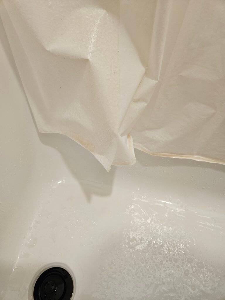 Close-up of a white shower curtain with visible pink mold stains at the bottom, above a wet tub.